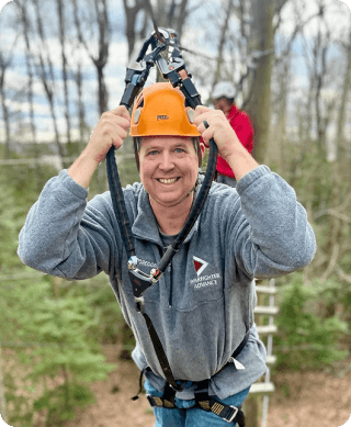 Smiling person wearing an orange helmet and zip line harness at an outdoor adventure course