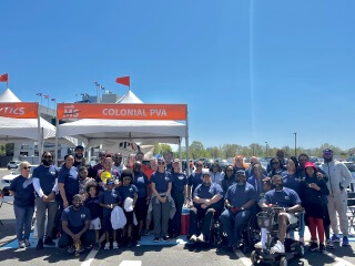Large group of Colonial PVA members and supporters, including a person in a wheelchair, posing outdoors at a sunny community event.
